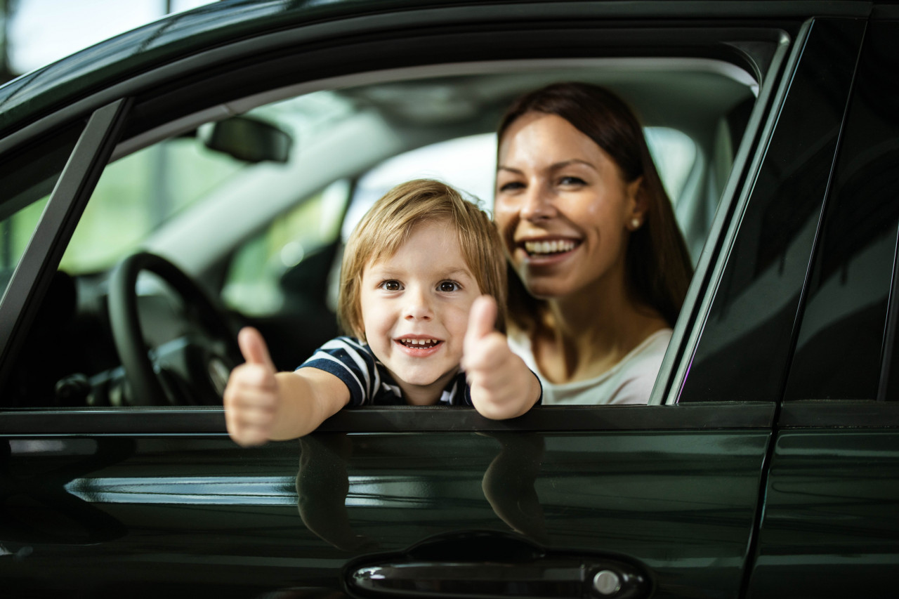 A little boy giving two thumbs up out the car window with his mom.