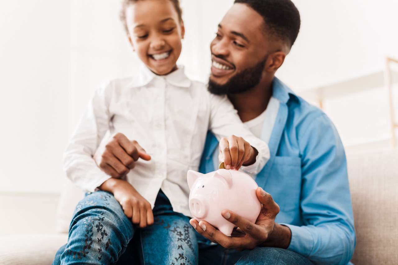 A young girl with her dad placing coins in her piggy bank.