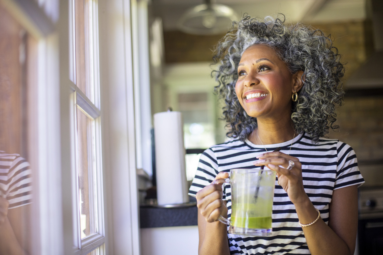 A woman drinking a green smoothie in her kitchen looking out the window.